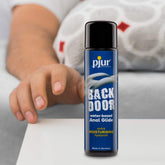 A bottle of Pjur Backdoor Comfort Water-Based Anal Glide rests on a surface. As a hand reaches for it, someone is partially visible in the background, possibly anticipating the smooth sensation provided by this Pjur lubricant.